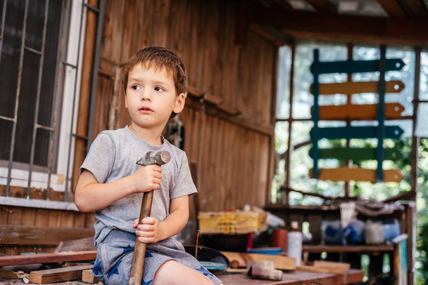 Little boy hammers nails with a hammer in a wooden board in the carpenters workshop - Zdjęcie, obraz