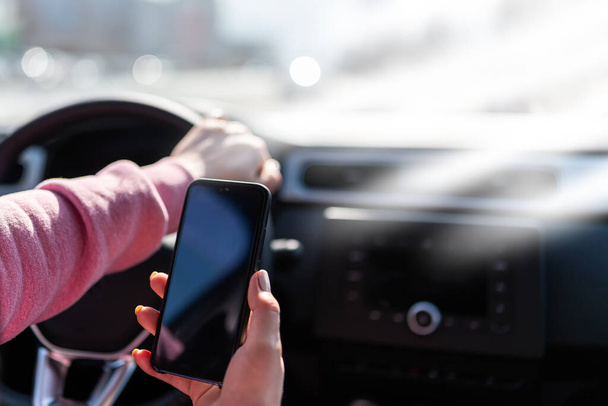 Hand houdt de mobiele telefoon tegen de achtergrond van het stuur van de auto. Afgeleide bestuurder met een mobiele telefoon. Telefoon met een blanco zwart scherm voor een inscriptie of afbeelding. Gevaar voor ongevallen  - Foto, afbeelding