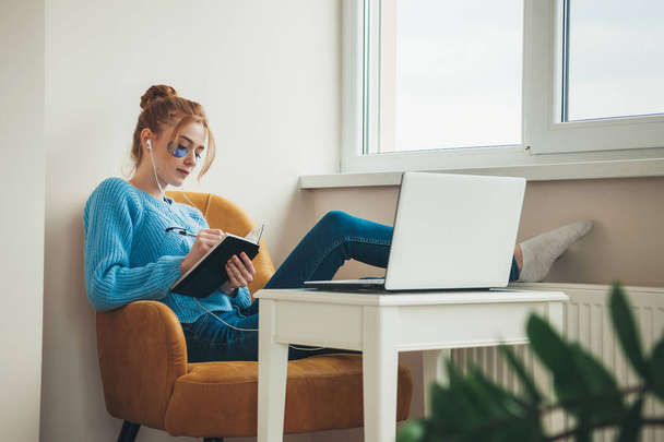 Ginger woman with freckles is wearing hydrogel eye patches while lying in armchair and doing homework at the laptop - Photo, Image