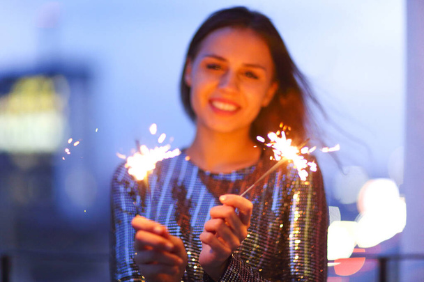 Entzückte junge Dame im Abendkleid lächelt und schwenkt brennende Wunderkerzen bei der nächtlichen Weihnachtsfeier - Foto, Bild