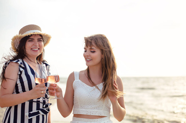Heureux adultes copines en tenue d'été souriant tout en dégustant du vin sur la plage de sable près de la mer - Photo, image