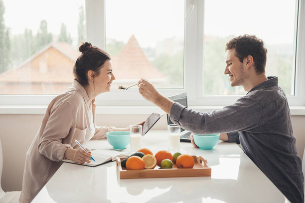 Caucasian careful man is feeding his girlfriend while working in the kitchen together on laptop - Photo, Image
