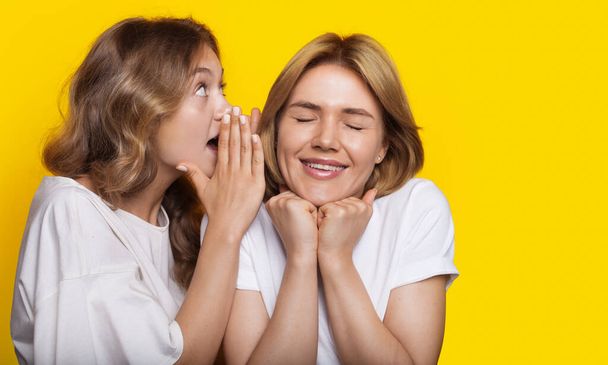 Blonde woman is whispering to her friend something while posing on a yellow studio wall - Photo, image