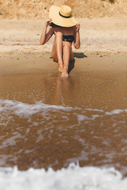 Mulher bonita e elegante em chapéu sentado na praia de areia em ondas do mar, banhos de sol e relaxante. Férias. Ajuste sexy jovem fêmea em maiô e chapéu de palha desfrutando de férias na ilha tropical - Foto, Imagem