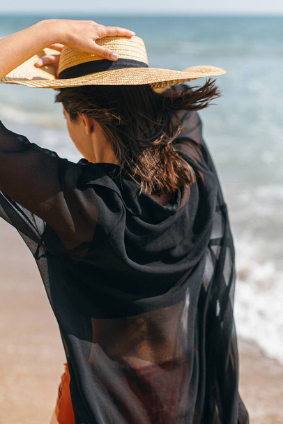 Beautiful stylish woman in hat walking on windy beach at sea waves. Summer vacation. Fit sexy young female in light shirt and straw hat relaxing on tropical island - Photo, Image