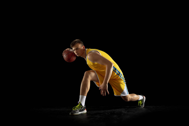 One young man, basketball player with a ball training isolated on dark black studio background. Advertising concept. Fit Caucasian athlete practicing with ball. - Foto, Imagem
