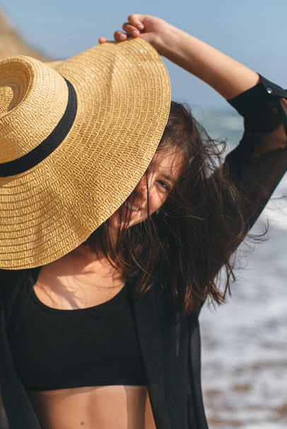 Beautiful happy woman with windy hair relaxing and smiling under hat on sandy beach at sea. Summer vacation.Stylish fit young female in light shirt and straw hat relaxing on tropical island - Фото, изображение