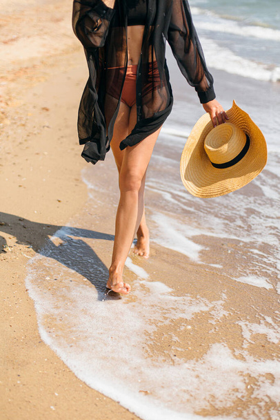 Carefree woman holding hat and walking on sunny beach at sea waves, close up. Stylish young fit female with straw hat in hands relaxing on tropical shore. Summer vacation - Photo, image
