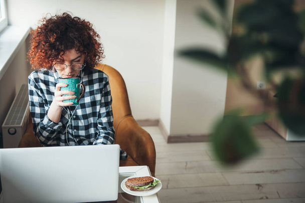Curly woman is drinking a coffee with burger while working at a laptop from home - Photo, Image