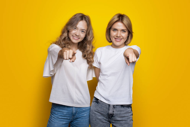Светловолосые сестры показывают на камеру, улыбаясь на желтой стене студии в футболке и джинсах. - Фото, изображение