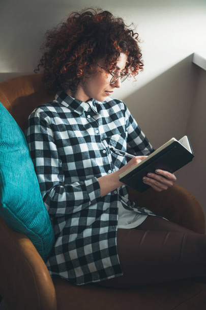 Imádnivaló, göndör hajú nő ír egy könyvbe, miközben otthon fekszik egy karosszékben szemüvegben. - Fotó, kép
