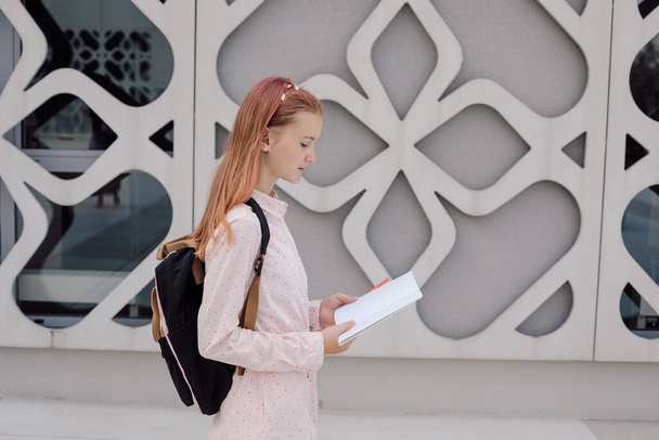 Teenage girl schoolgirl on background of concrete building with backpack on back with book , copy space - Photo, Image