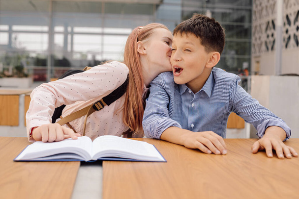 Μια έφηβη μαθήτρια ψιθυρίζει κάτι στο αυτί ενός αγοριού ή τον φιλάει.. - Φωτογραφία, εικόνα