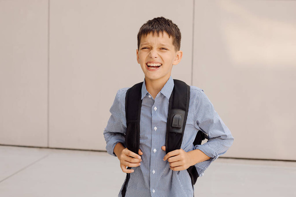 Junge Teenager 11 Jahre alter Schüler blickt in die Kamera auf grauem Hintergrund mit Rucksack lächelnd. Im karierten Hemd gekleidet - Foto, Bild
