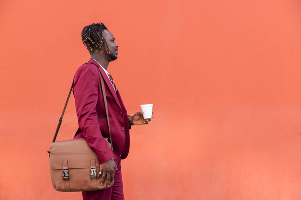 elegant african businessman in a suit with briefcase holding a coffee in his hand in front of a red background, copy space for text - Foto, afbeelding