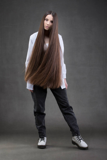 Ένα όμορφο κορίτσι με πολύ μακριά μαλλιά σε πλήρη ανάπτυξη σε γκρι φόντο. - Φωτογραφία, εικόνα