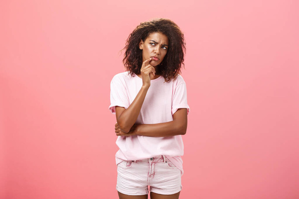 Suspicious displeased dark-skinned young curly-haired woman in trendy t-shirt and shorts saying hmm touching chin with finger frowning and staring left doubtful and unsure having concern - Photo, Image