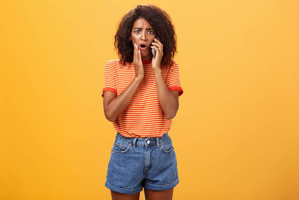 Woman receiving disturbing call feeling emapthy and sorry for poor friend getting in trouble holding cellphone near ear touching cheek concerned frowning standing sad, worried over orange background - Foto, imagen