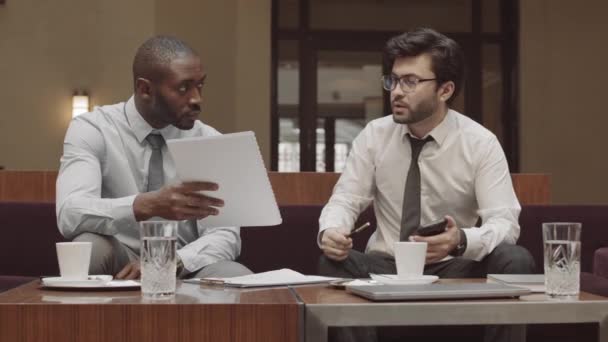 Orta uzunlukta iki genç erkek iş ortağı şık bir restoranda kanepede oturuyor, koyu saçlı CEO sözleşme anlaşmasını okuyor ve reddediyor, Afrikalı müdür ayrılıyor. - Video, Çekim