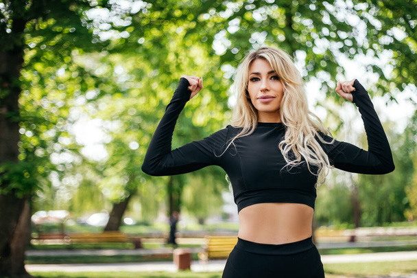 Woman standing posing showing body biceps abdominal muscles outside green park morning looking away. Athletic lady dressed fitness sportwear clothes. Healthy lifestyle, sport recreation. Copy space - Photo, Image