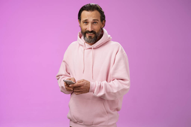 Stílusos jóképű felnőtt szakállas srác 40-es divatos rózsaszín kapucnis gazdaság smartphone SMS megjelenés kamera zavartalan hűvös magabiztos zavart játszik félelmetes új app játék, lila háttér - Fotó, kép