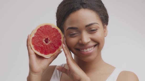Vitaminas para cuidado de la piel y belleza. Mujer afroamericana bastante alegre sosteniendo media toronja cerca de su cara - Imágenes, Vídeo