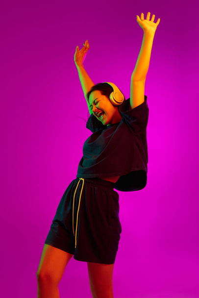 Studio shot of young pretty caucasian girl with headphones dancing to the music isolated over pink background in neon lights - 写真・画像