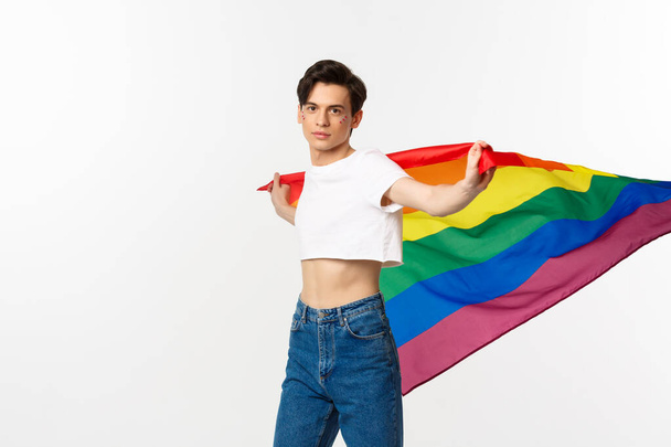 Права людини та концепція спільноти lgbtq. Ззовні і гордий гей чоловік махає веселковим прапором і впевнено дивиться на камеру, стоїть на вершині врожаю і джинси на білому тлі
 - Фото, зображення