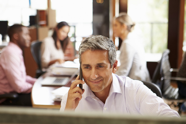 Businessman On Phone At Desk With Meeting In Background - Photo, Image