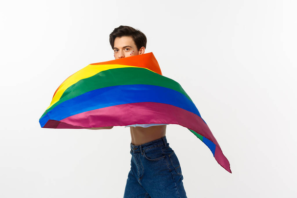 Mensenrechten en het concept van de gemeenschap. Jonge homo met glitter op het gezicht, zwaaiend lgbtq vlag met trots, staande over witte achtergrond - Foto, afbeelding