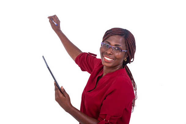 una hermosa mujer de negocios en gafas de pie en una habitación escribiendo con un bolígrafo en una pizarra mientras sostiene una tableta y mirando a la cámara mientras sonríe. - Foto, imagen