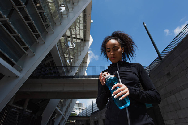 Low angle view van Afrikaanse Amerikaanse sportvrouw op zoek naar sport fles op stedelijke straat  - Foto, afbeelding