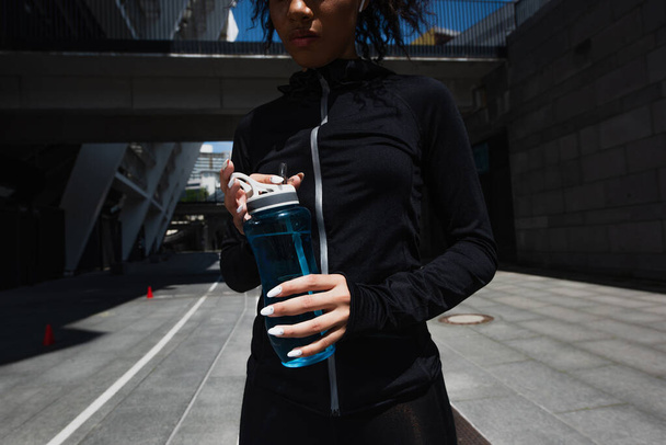 Gekropte weergave van Afrikaanse Amerikaanse sportvrouw in oortelefoon houden sport fles buiten  - Foto, afbeelding