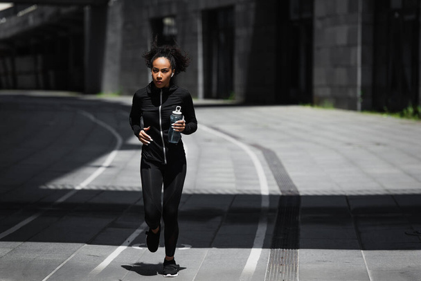 African american runner with sports bottle listening music in earphones on urban street  - Photo, Image