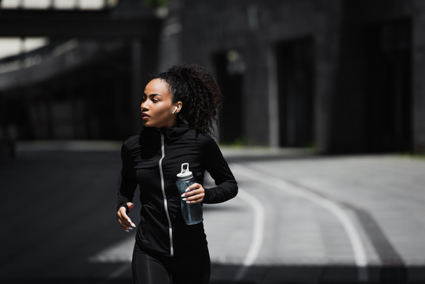 Afrikanischer Läufer mit drahtlosem Kopfhörer hält Sportflasche im Freien  - Foto, Bild