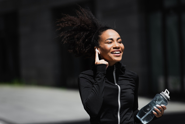 Corredor afroamericano positivo con botella de deporte usando auriculares al aire libre  - Foto, Imagen