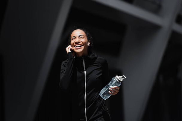 Smiling african american sportswoman in earphones holding sports bottle outdoors  - Фото, изображение