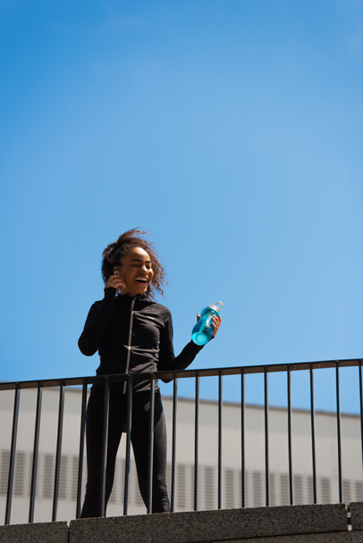 Vista de bajo ángulo de la deportista afroamericana escuchando música en auriculares y sosteniendo la botella de deportes en el puente  - Foto, imagen