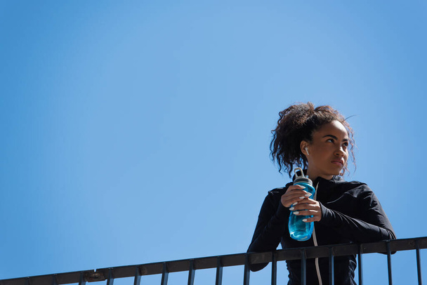 Low angle view of african american sportswoman in earphone holding sports bottle with blue sky at background - Foto, Bild
