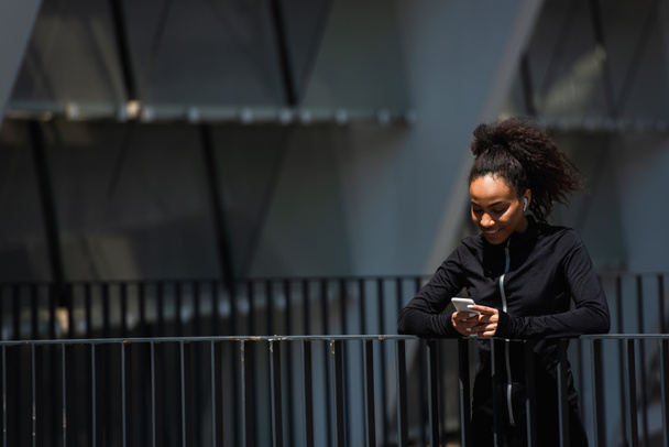 Smiling african american sportswoman in earphone using cellphone on urban street  - Foto, Imagen