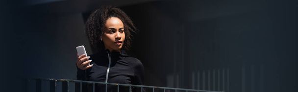 Afro-Amerikaanse sportvrouw in oortelefoon houden mobiele telefoon buiten, banner  - Foto, afbeelding