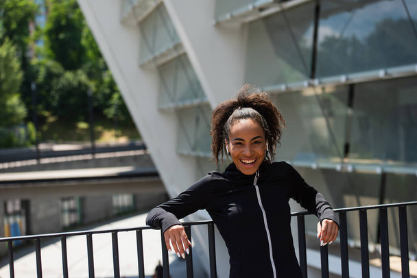 Lächelnde afrikanisch-amerikanische Sportlerin mit Kopfhörern, die in der Nähe einer Brücke auf der städtischen Straße steht  - Foto, Bild