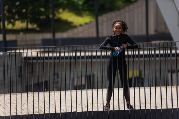 Positive african american sportswoman holding sports bottle and looking at camera on bridge  - Φωτογραφία, εικόνα