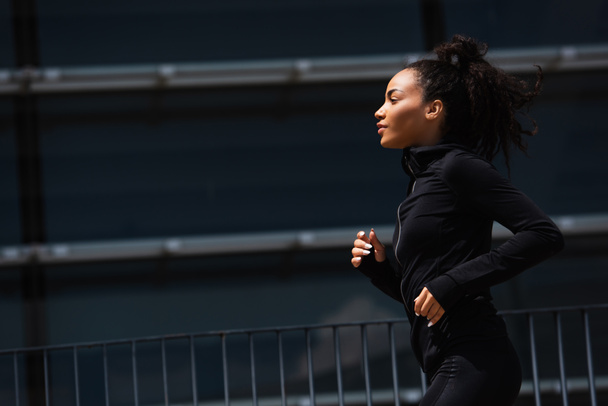 Boční pohled na africký americký běžec ve sportovní bundě na městské ulici  - Fotografie, Obrázek