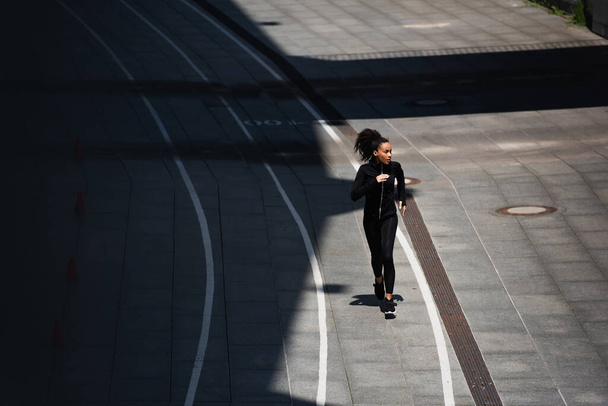 Wysoki kąt widzenia afrykańskiej amerykanki w czarnej odzieży sportowej biegającej na torze  - Zdjęcie, obraz