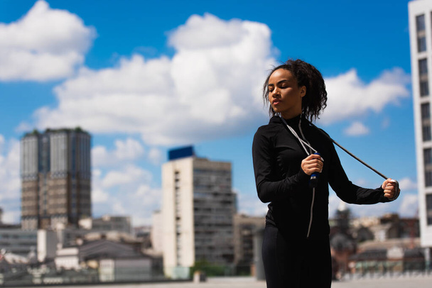 Young african american sportswoman with closed eyes holding skipping rope on urban street  - Foto, afbeelding