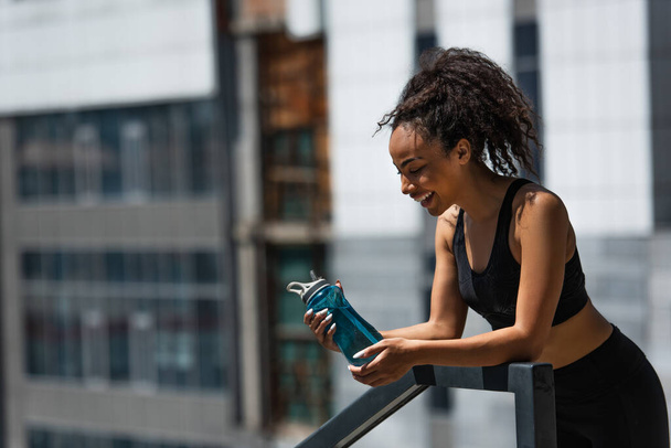 Side view of happy african american sportswoman looking at sports bottle on urban street  - Фото, изображение