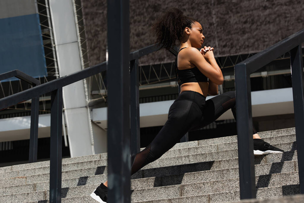 Side view of african american sportswoman doing lunges on stairs on urban street  - Fotó, kép