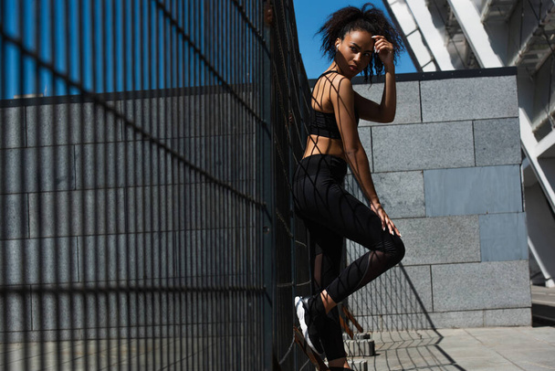 Mujer afroamericana en ropa deportiva y auricular de pie cerca de la valla en la calle urbana  - Foto, imagen