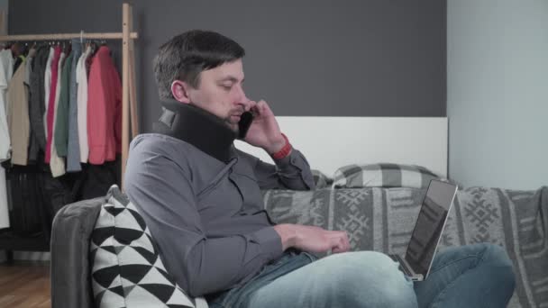 Boyunluk takan adam tele-telden çalışıyor. Köpüklü boyun tasması takmış yaralı bir erkek telefonda dizüstü bilgisayarla konuşuyor kanepede oturuyor ve boynu ağrıyor. - Video, Çekim